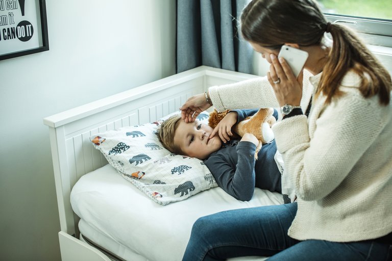Mor sitter på sengekanten til sin sønn og snakker i mobiltelefon