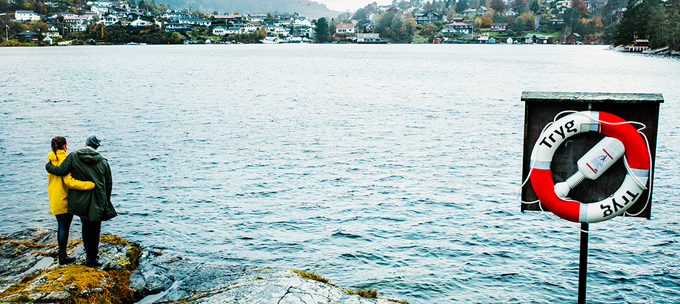 Tryg sin livbøye henger på et stativ ved sjøen. Et par omfavner hverandre.