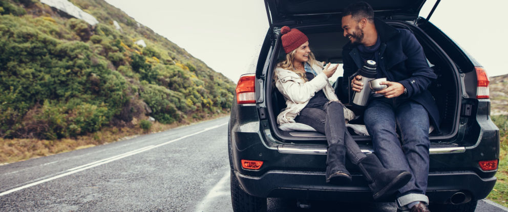Mann og kvinne sitter i åpent bagasjerom på bil og drikker kaffe