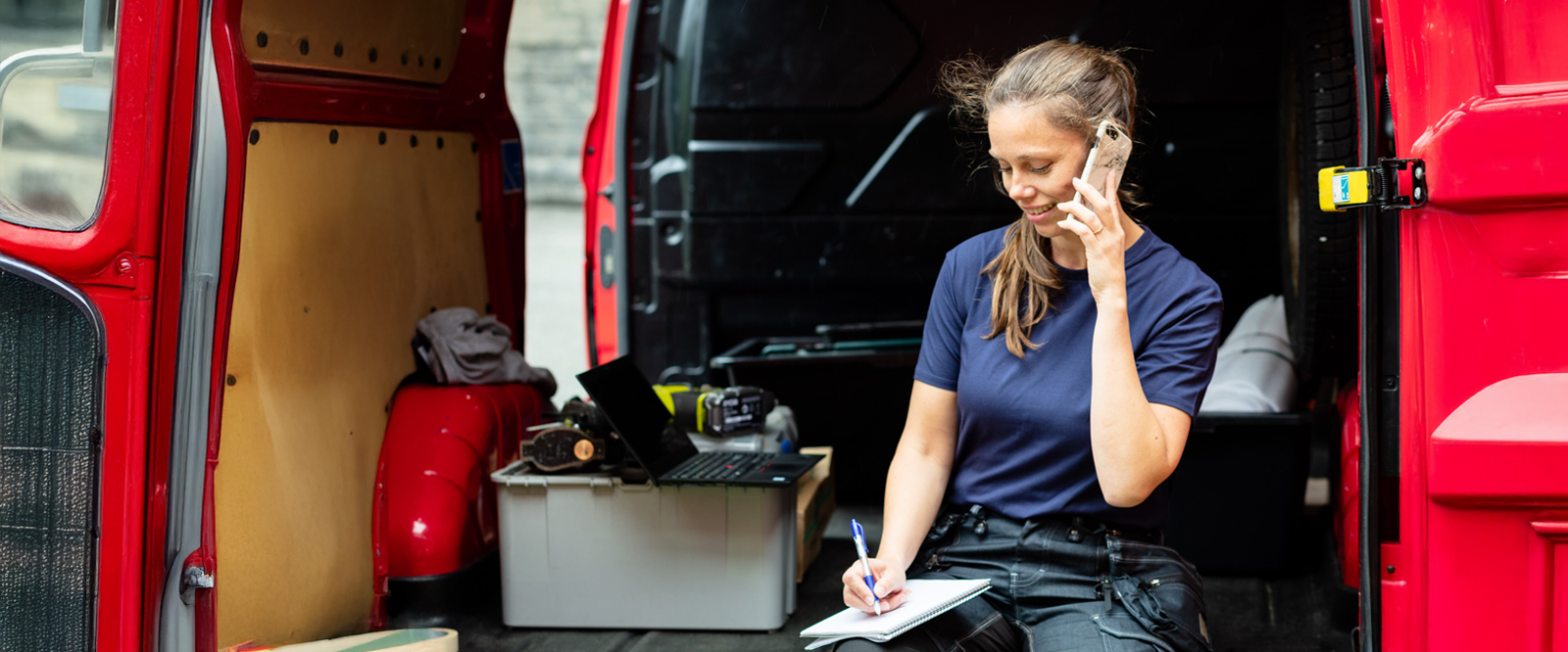 Kvinne sitter i arbeidsbilen og snakker i telefonen og noterer