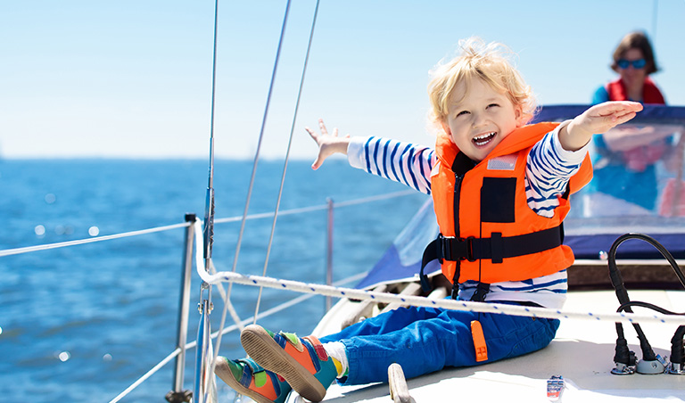 Liten gutt i redningsvest sitter på dekk på en seilbåt og smiler