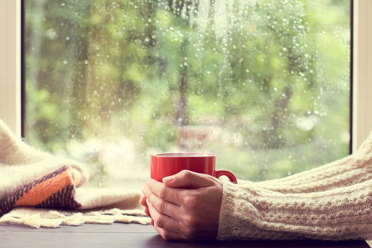 En person sitter med en kopp mellom hendene og ser ut på regnfult høstvær