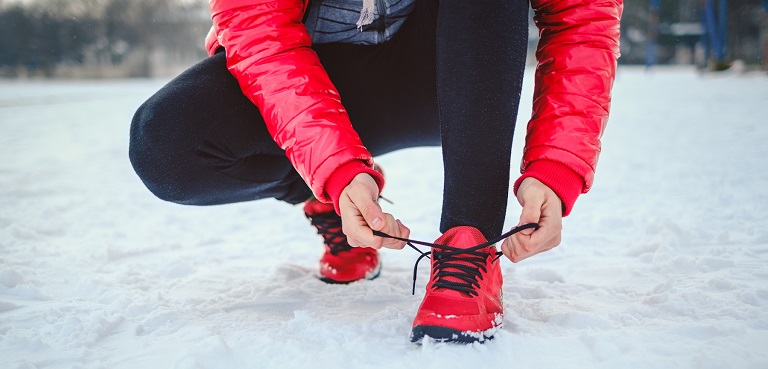 Ute i snø, står en person med rød boblejakke og røde joggesko og knyter ene skolissen.