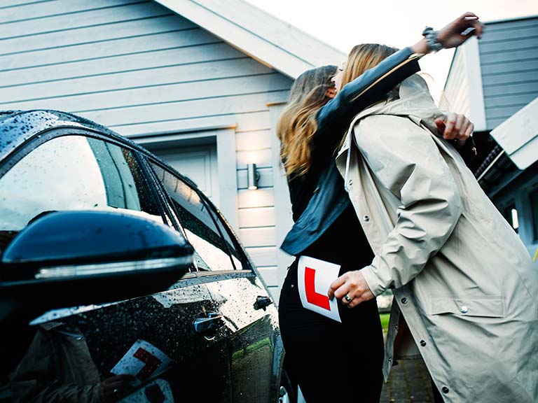 En datter klemmer sin mor ved siden av en bil. Moren holder et L-skilt i hånden.