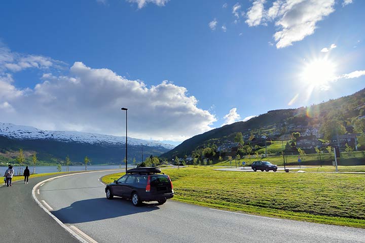 Bil på vei gjennom en norsk bygd med hvitkledte sommerfjell i bakgrunnen